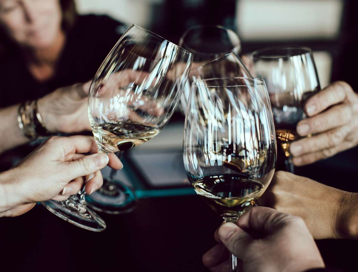 Group of people clinking glasses in a celebratory toast with white and amber wine at a dimly lit gathering.
