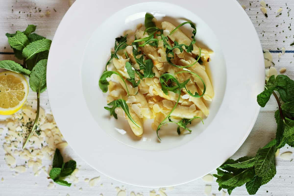 White asparagus salad garnished with arugula, almond flakes, and lemon, surrounded by fresh mint on a white cloth.