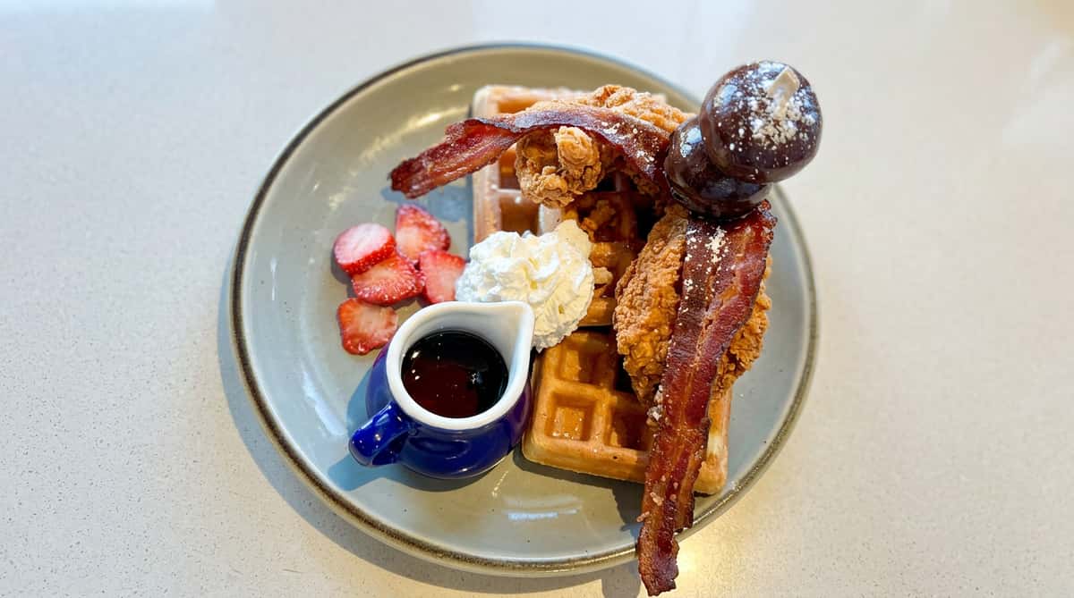 Chicken and waffles dish with crispy bacon, strawberries, whipped cream, a side of syrup, and chocolate covered grapes.