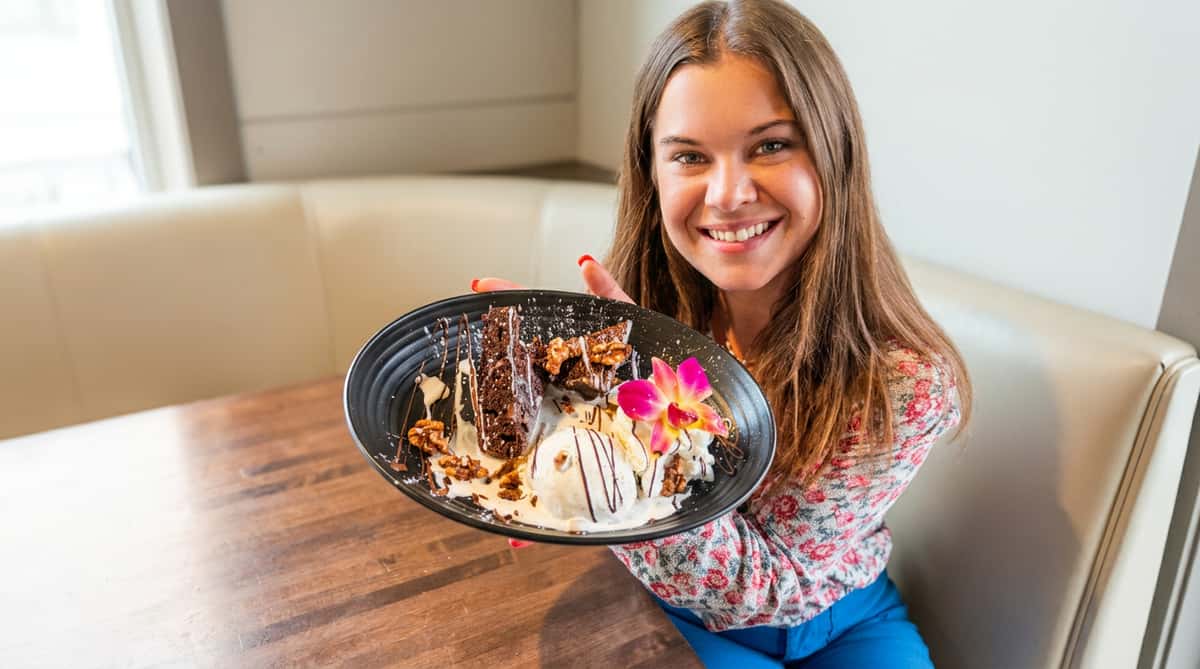 A smiling woman holds a plate with chocolate brownies, ice cream, walnuts, and a pink orchid.