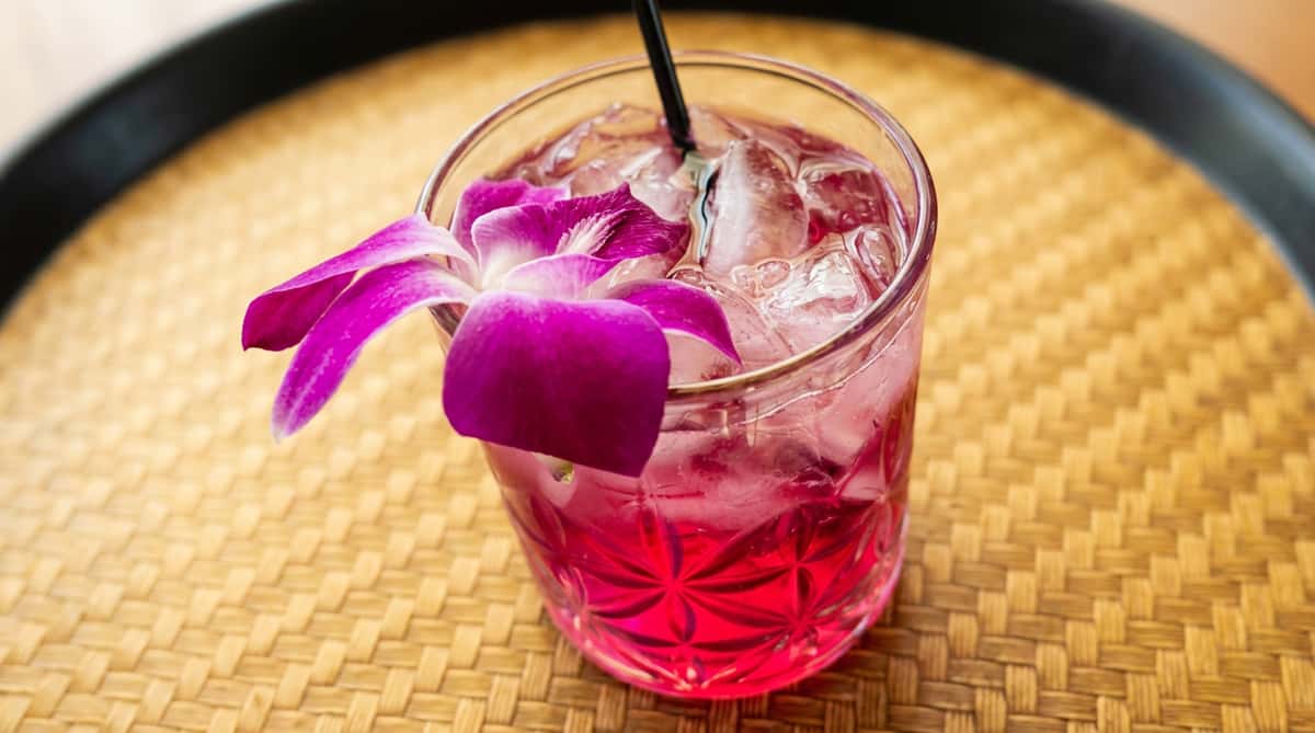 Pink cocktail with ice cubes, garnished with a bright fuchsia orchid, on a woven serving tray.