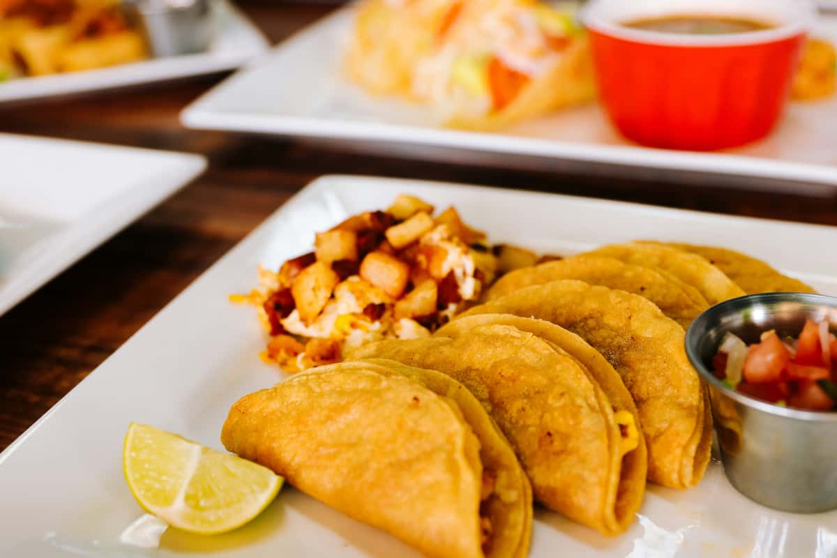Crispy golden tacos served with a side of seasoned potatoes, fresh pico de gallo, and a lime wedge on a white plate — a delicious Mexican dish perfect for a casual dining experience.