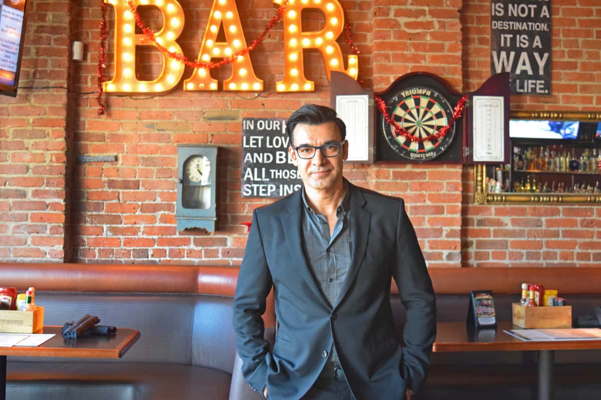 Professional standing inside a stylish brick-walled bar featuring illuminated marquee signage, dartboards, and a warm neighborhood pub atmosphere.