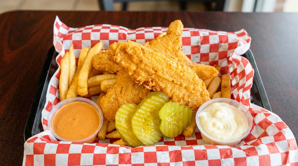 Fish and chips with pickles, a red sauce, and white sauce, served in a basket with red and white checkered paper.