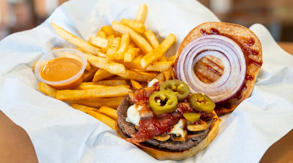 An open hamburger with beef patty, bacon, jalapenos, mushrooms, and BBQ sauce served with a pile of golden fries and dipping sauce in a white basket.