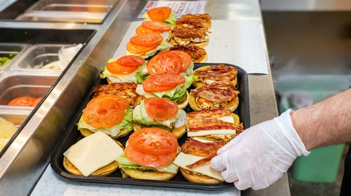 An assembly line of burgers and bacon sandwiches on a tray, topped with lettuce, tomato, and cheese.