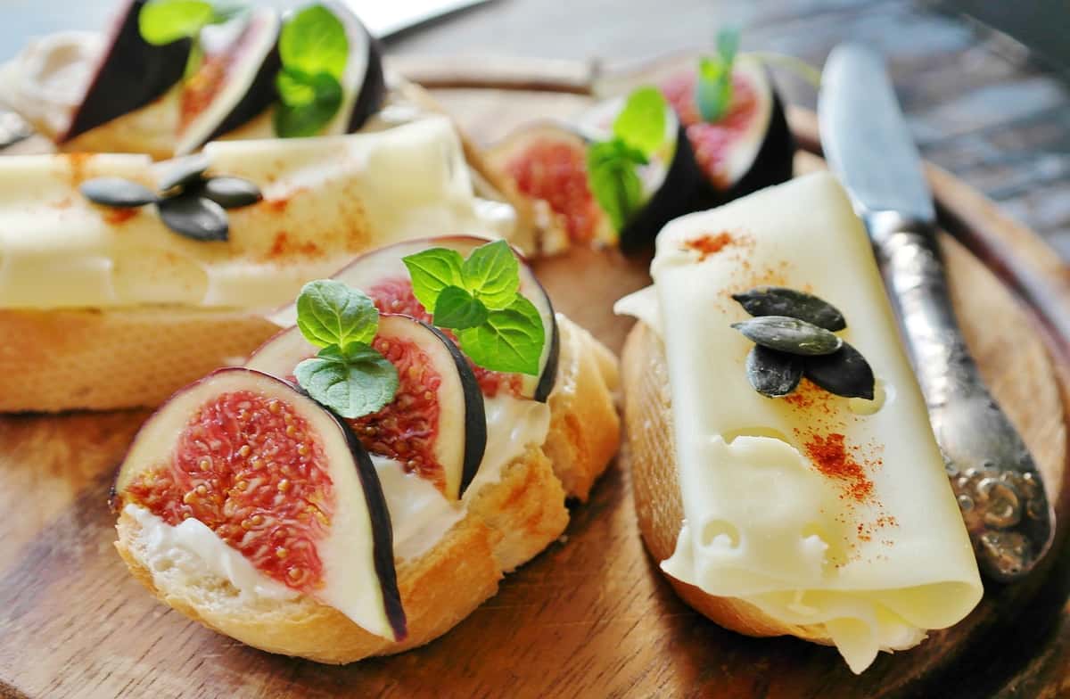 Gourmet baguette slices topped with figs, soft cheese, fresh mint, pumpkin seeds, and paprika on a rustic wooden board.