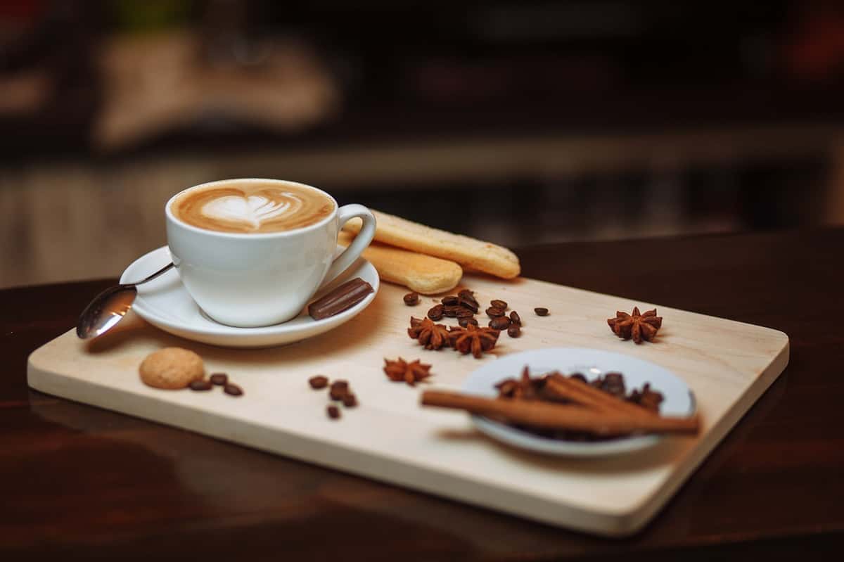 Latte with heart-shaped foam art served on a wooden board with cookies, cinnamon sticks, star anise, and coffee beans.