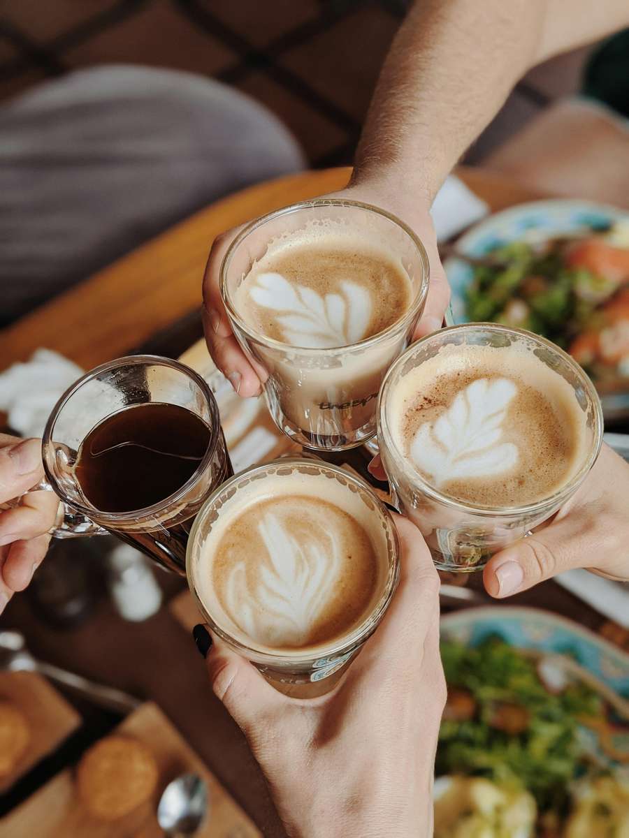 Four hands holding up glasses of coffee with latte art and black coffee in a toast gesture over a table with food.