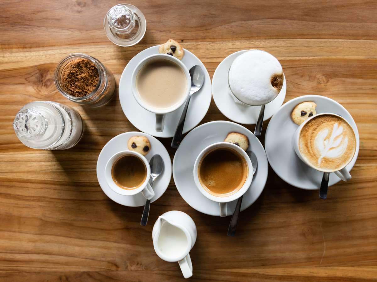 Assorted coffee drinks on a wooden table with cookies, sugar, milk, and spoons arranged neatly for a cozy café setting.