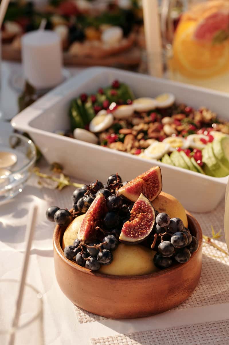A wooden bowl of fresh figs, grapes, and apples on a sunlit table with a vibrant salad and citrus drink in the background.