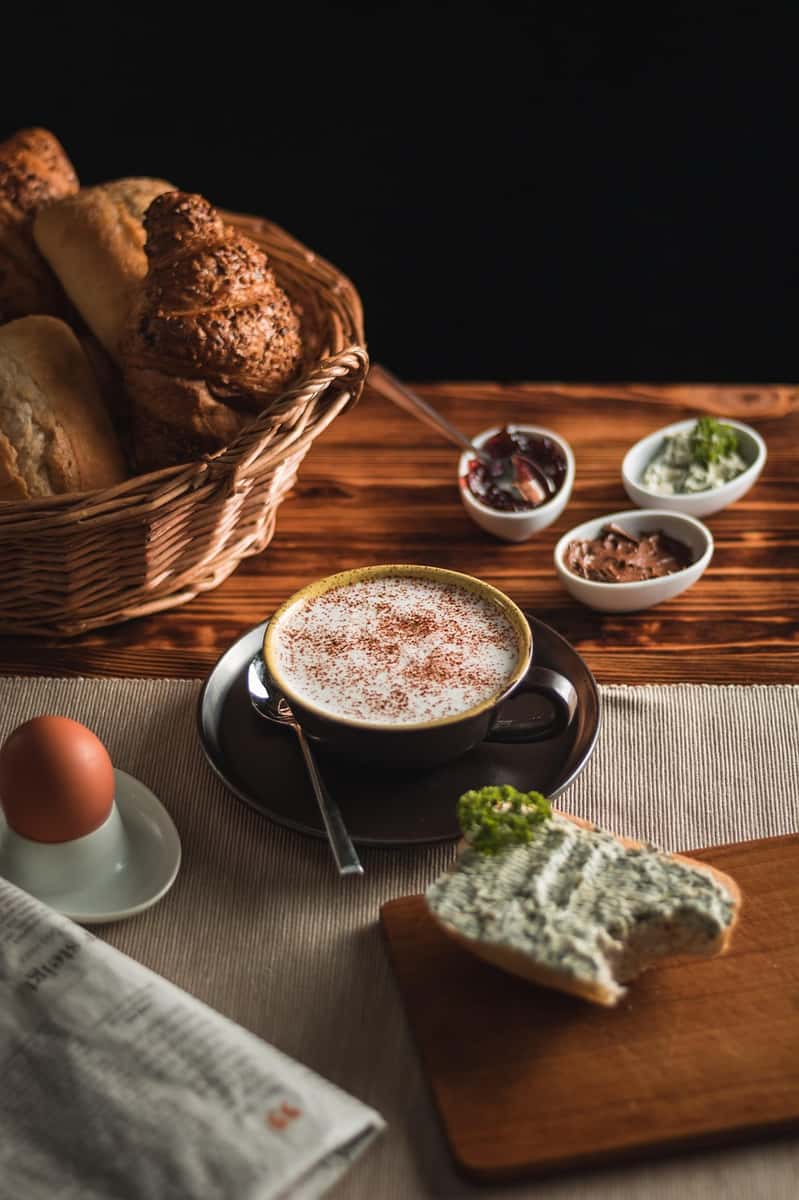 Foamy cappuccino with cinnamon, fresh bread, spreads, a soft-boiled egg, and a herbed cream cheese sandwich on a cozy breakfast table.