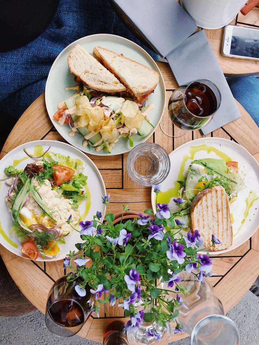 Outdoor café table with artisan sandwiches and vibrant salad plates, served with crisp potato chips, fresh greens, cherry tomatoes, and creamy dressings, paired with glasses of iced tea and a centerpiece of purple pansies.
