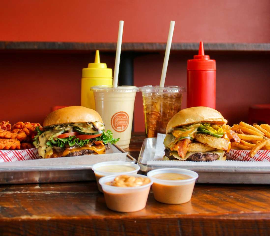 Social Burger meal featuring two gourmet burgers with crispy fries, chicken bites, dipping sauces, soft drinks, and a signature milkshake on a restaurant table.
