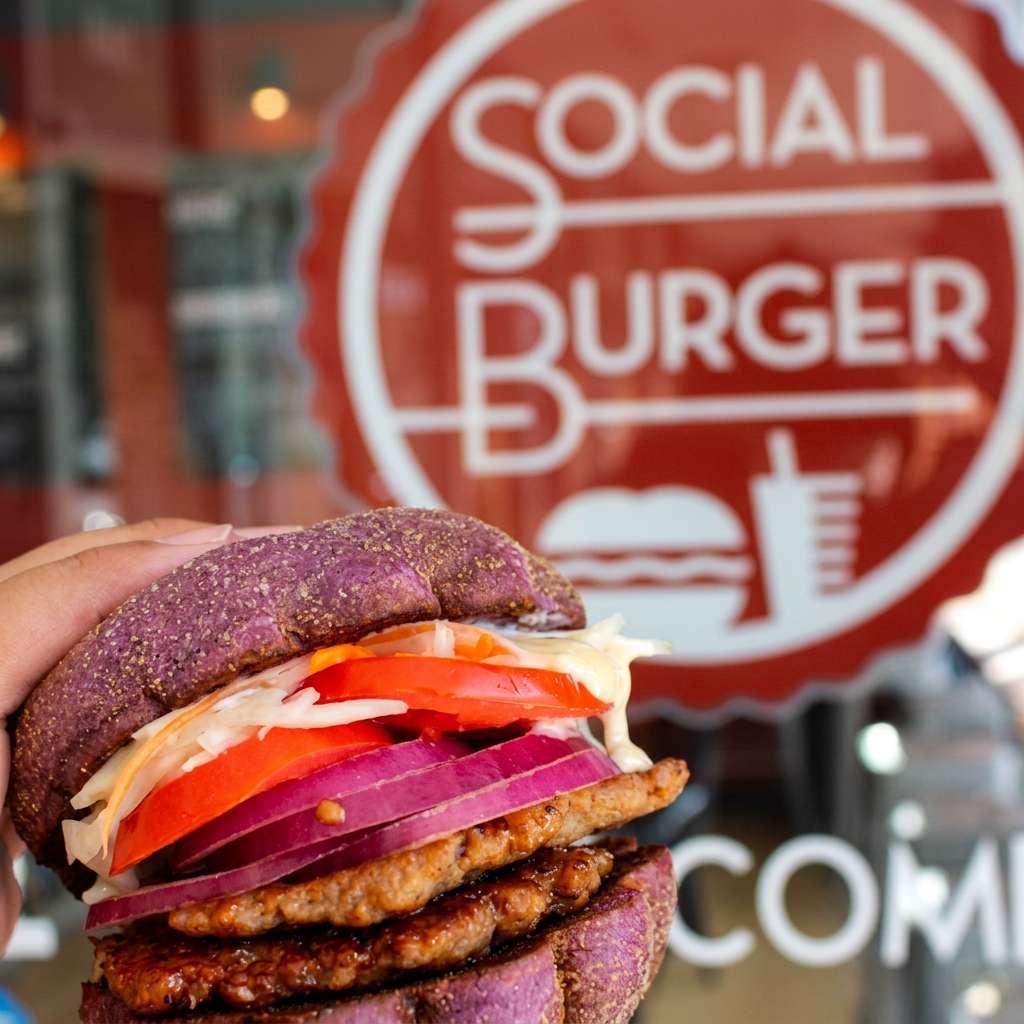 Hand holding a Social Burger sandwich on a purple bun with grilled patties, fresh tomatoes, red onions, and shredded cabbage in front of the restaurant storefront.