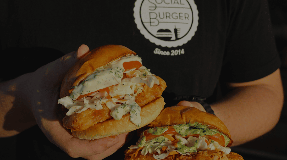 Two crispy fried chicken sandwiches topped with creamy herb sauce, fresh coleslaw, and tomatoes, held by a Social Burger team member wearing a branded restaurant shirt.