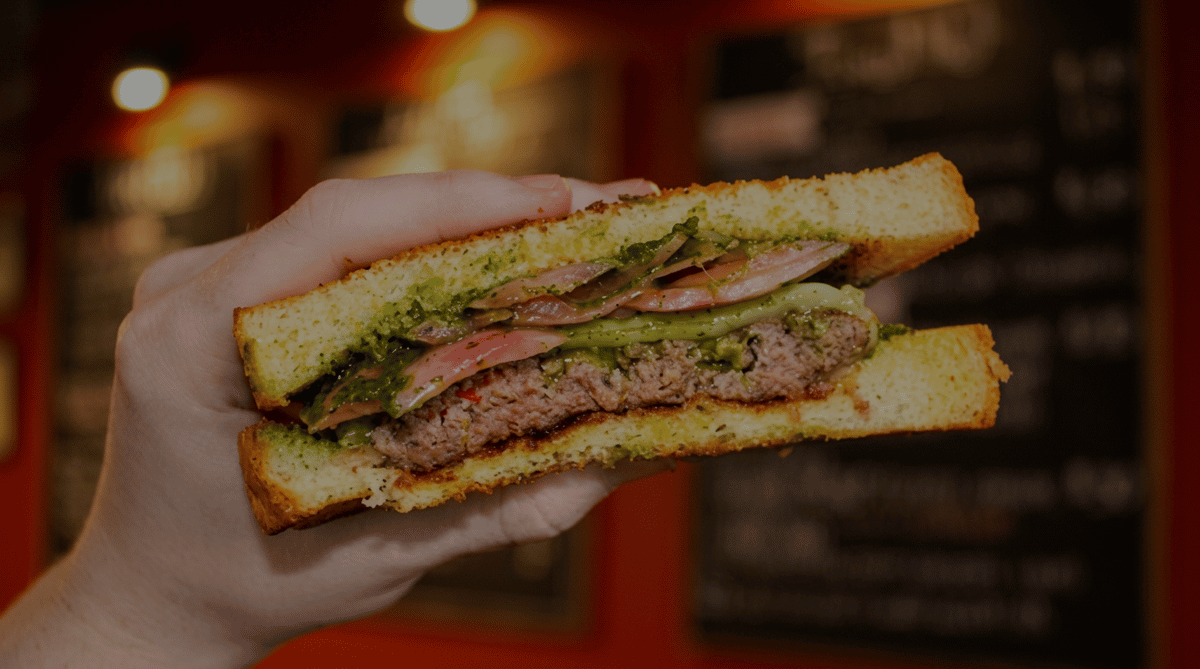 Hand holding a Social Burger garlic toast burger with a juicy beef patty, avocado, pickled red onions, and herb sauce on toasted bread inside the restaurant.