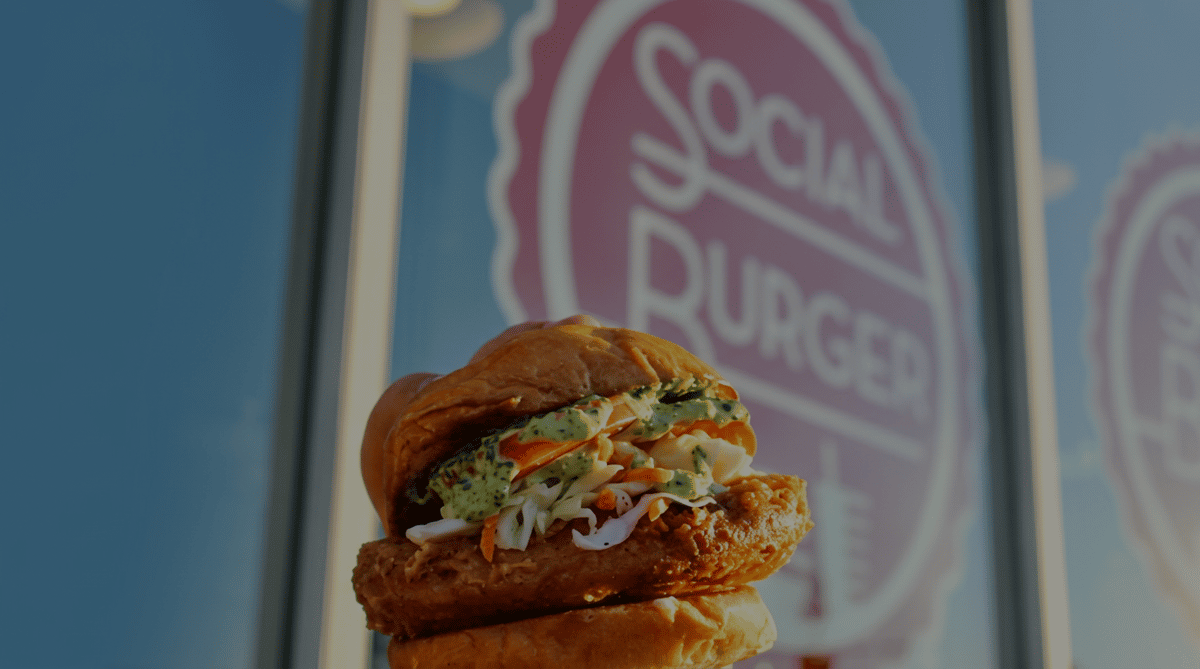 Crispy fried chicken sandwich topped with creamy herb sauce, fresh coleslaw, and tomatoes in front of Social Burger restaurant signage.