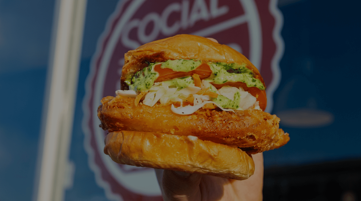 Crispy fried chicken sandwich topped with creamy herb sauce, fresh coleslaw, and sliced tomatoes, held in front of the Social Burger restaurant storefront.