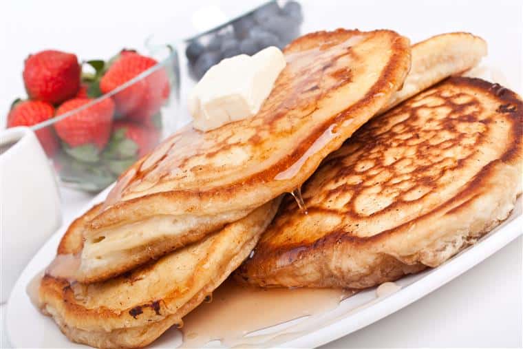 stack of pancakes topped with butter and syrup with blueberries and strawberries on the side