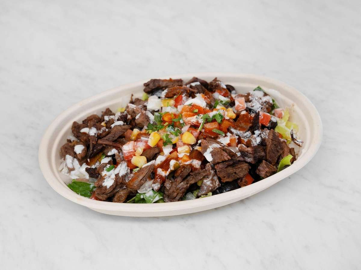 Bowl of grilled carne asada over a bed of lettuce and rice, topped with pico de gallo, corn, and drizzled with crema and salsa.