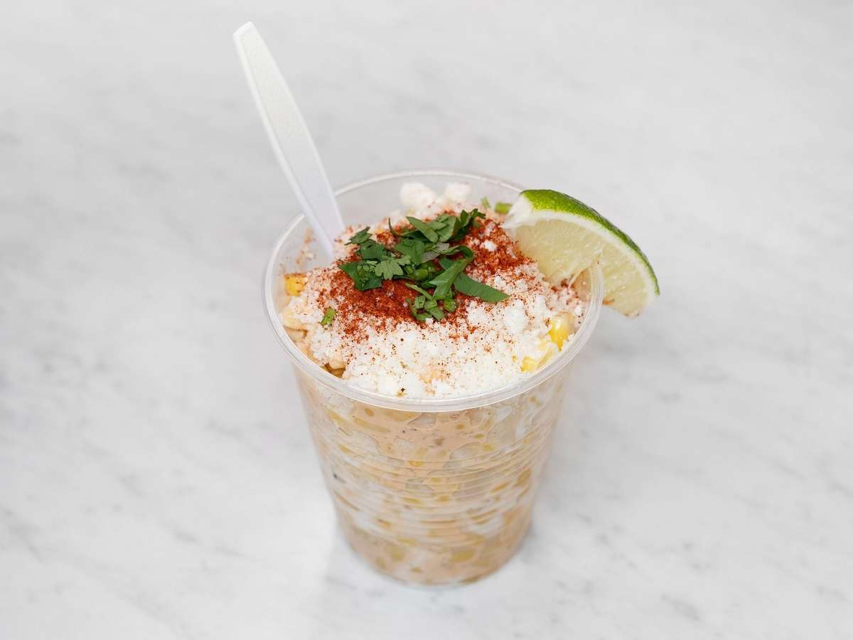 Cup of Mexican street corn topped with cotija cheese, chili powder, and chopped cilantro, garnished with a lime wedge on a marble surface.