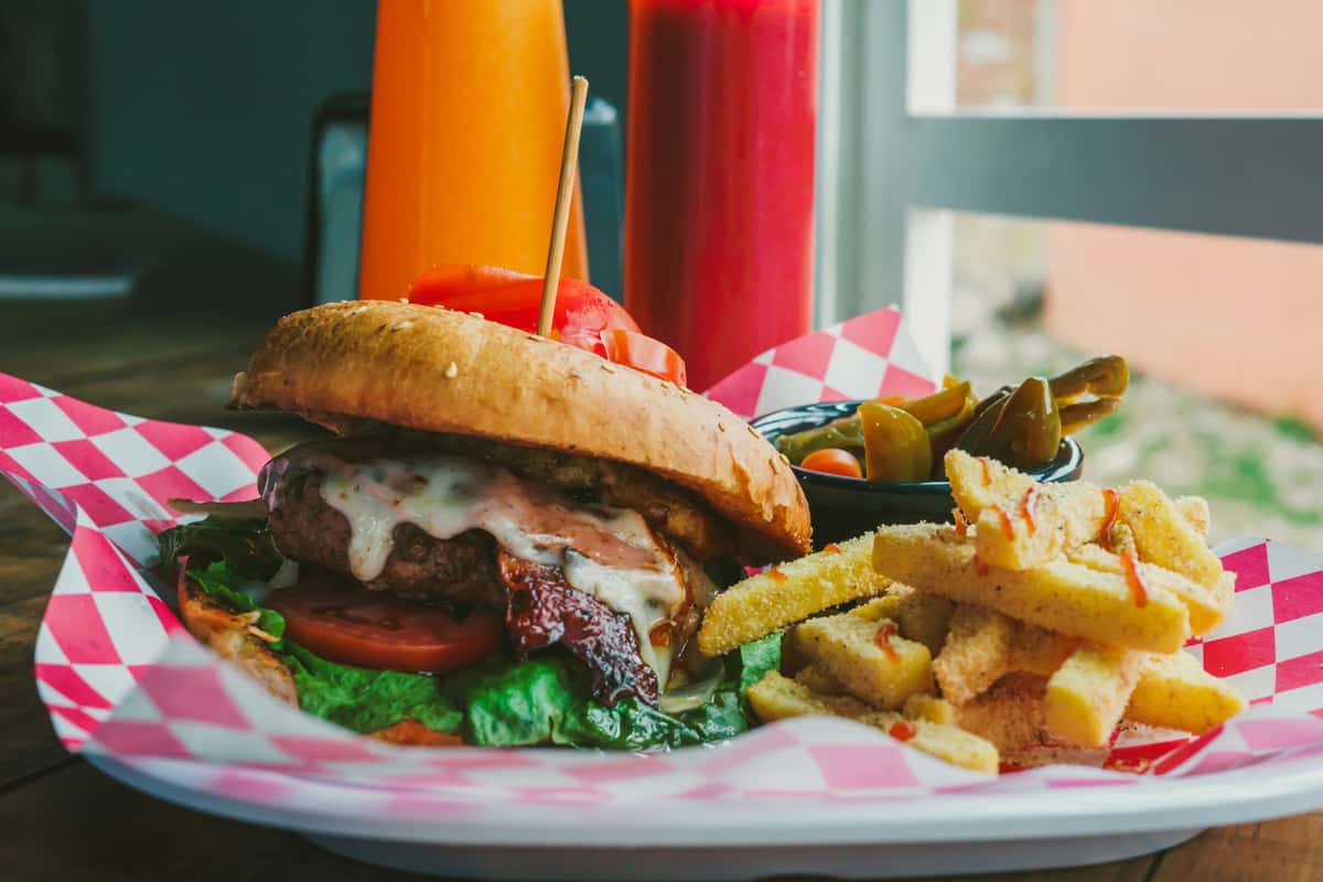 Juicy cheeseburger with lettuce, tomato, crispy bacon, and melted cheese on a toasted bun, served with thick-cut seasoned fries and a side of pickled jalapeños, presented in a red-and-white checkered tray with colorful drinks in the background.