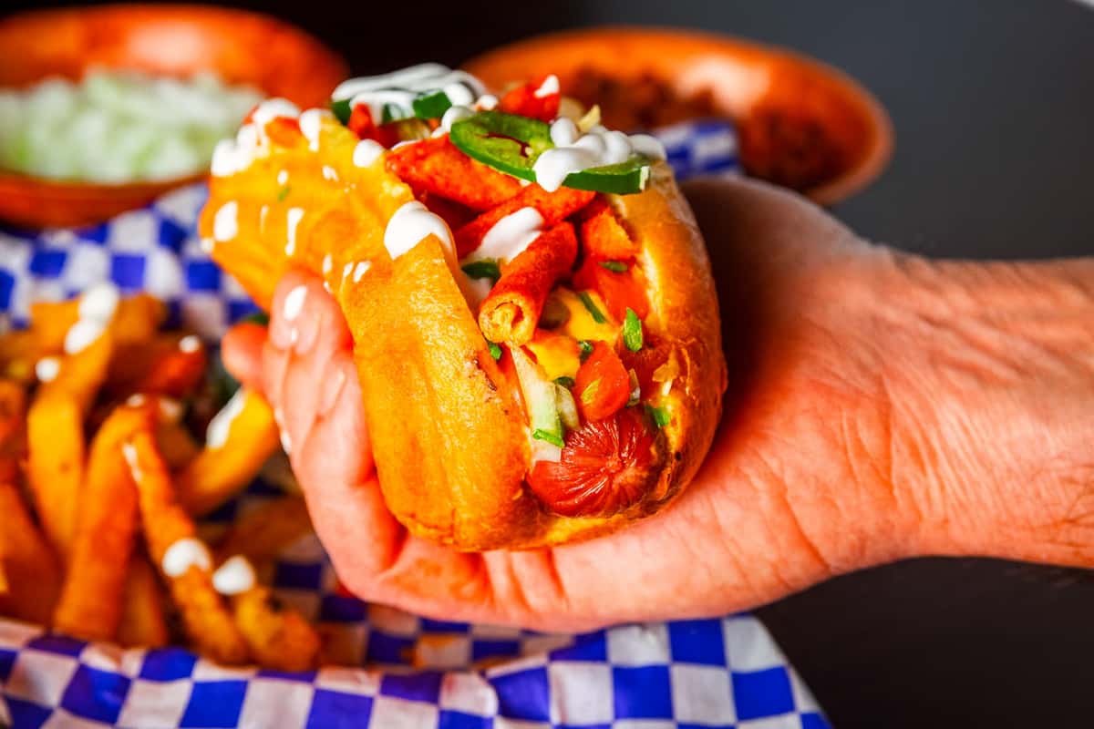 Close-up of a loaded hot dog topped with diced onions, jalapeño slices, crunchy rolled chips, melted cheese, and drizzled with sour cream, served in a toasted bun and held over a tray of seasoned fries.