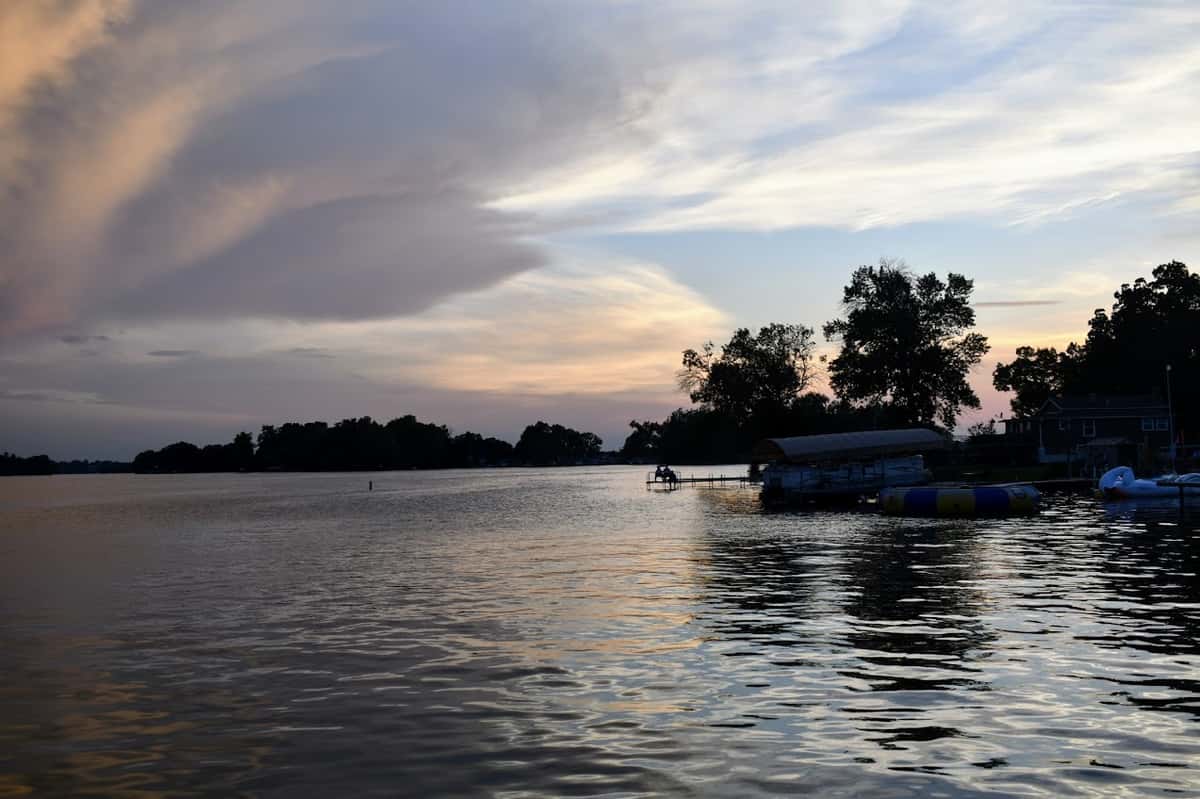 Scenic lakeside view at sunset with calm rippling water, silhouetted trees, and a dock with moored boats near a shoreline cottage under a colorful sky.