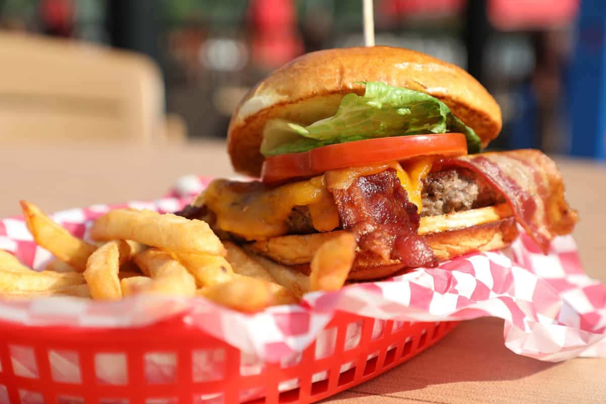 Juicy bacon cheeseburger stacked with cheddar cheese, crispy bacon, fresh tomato, and lettuce on a toasted bun, served with golden French fries in a red checkered basket for a classic American diner-style meal.