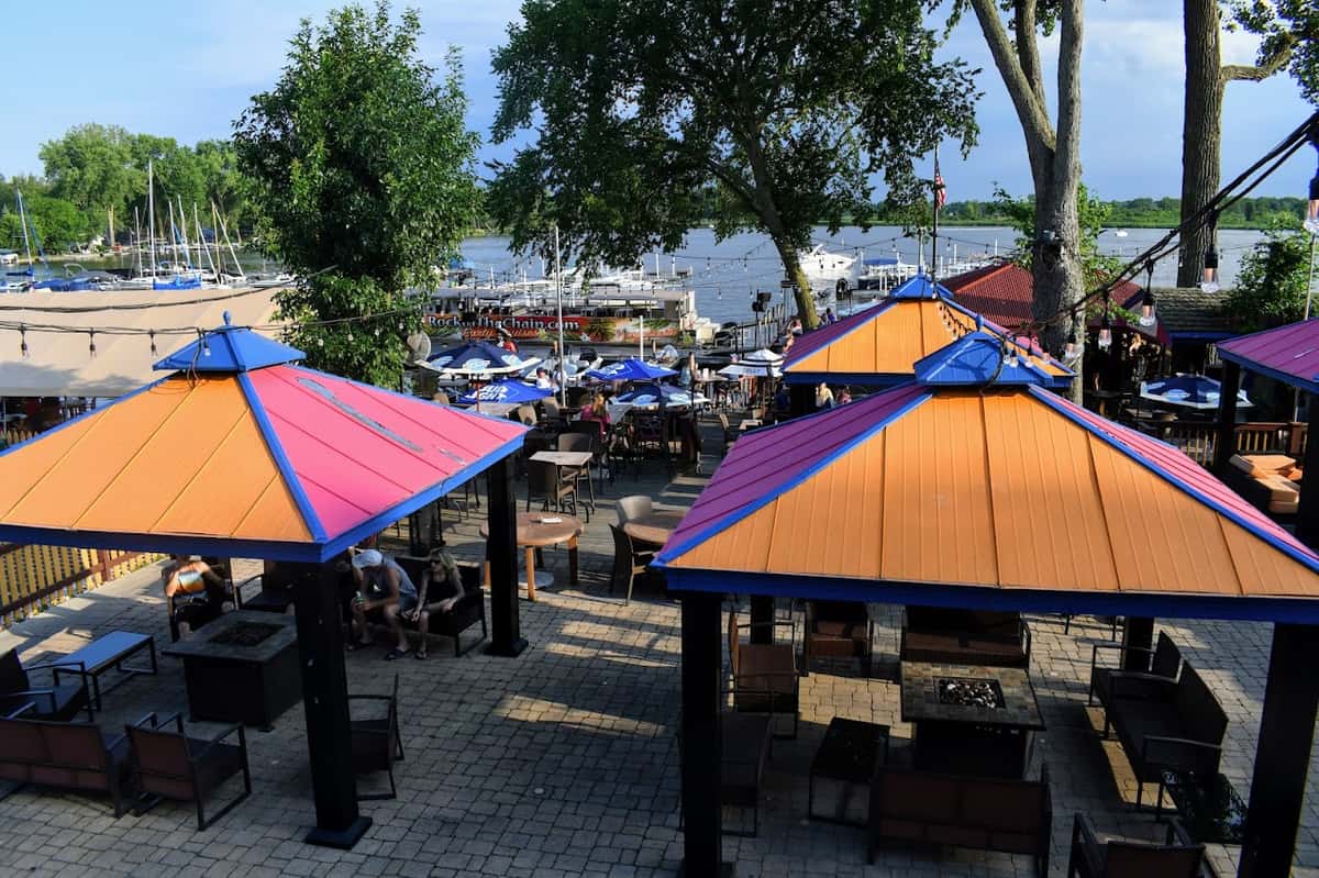 Outdoor dining patio with vibrant cabana-style umbrellas, lounge seating, and string lights, overlooking a lively marina filled with boats.