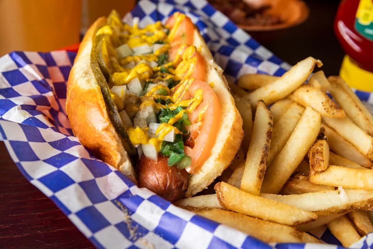 Classic Chicago-style hot dog topped with mustard, onions, tomatoes, relish, and a pickle spear, served in a soft bun with a side of golden French fries in a blue checkered basket.
