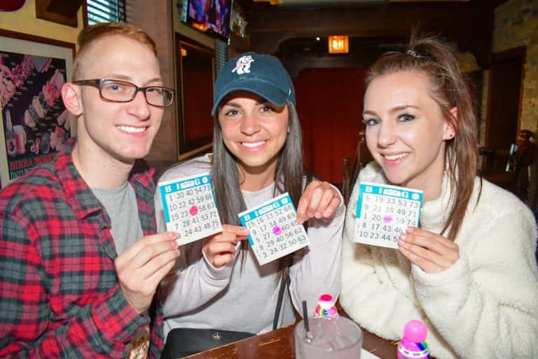 Three friends smiling and holding up bingo cards at a lively bar, with colorful bingo markers and drinks on the table, creating a fun game night atmosphere.