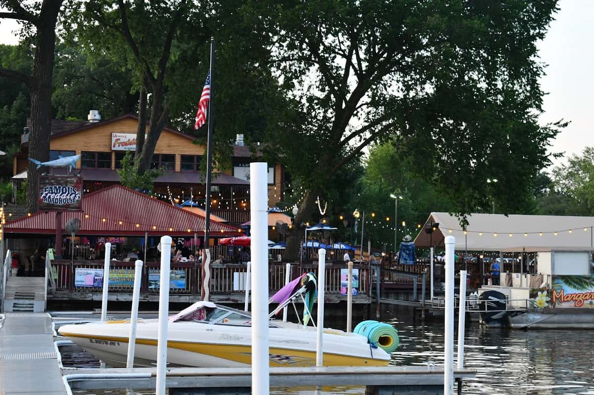 Waterfront restaurant and bar with outdoor seating, string lights, and colorful umbrellas, located along a marina with boats docked nearby, creating a lively lakeside dining atmosphere.