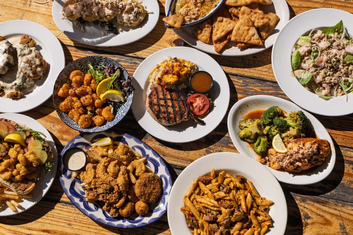 Overhead view of a rustic wooden table filled with assorted bar and grill dishes, including grilled steak with mashed potatoes, fried chicken tenders, crispy wings, loaded pasta, roasted broccoli, fresh salad, and appetizers, showcasing a hearty local American bar and grill menu.