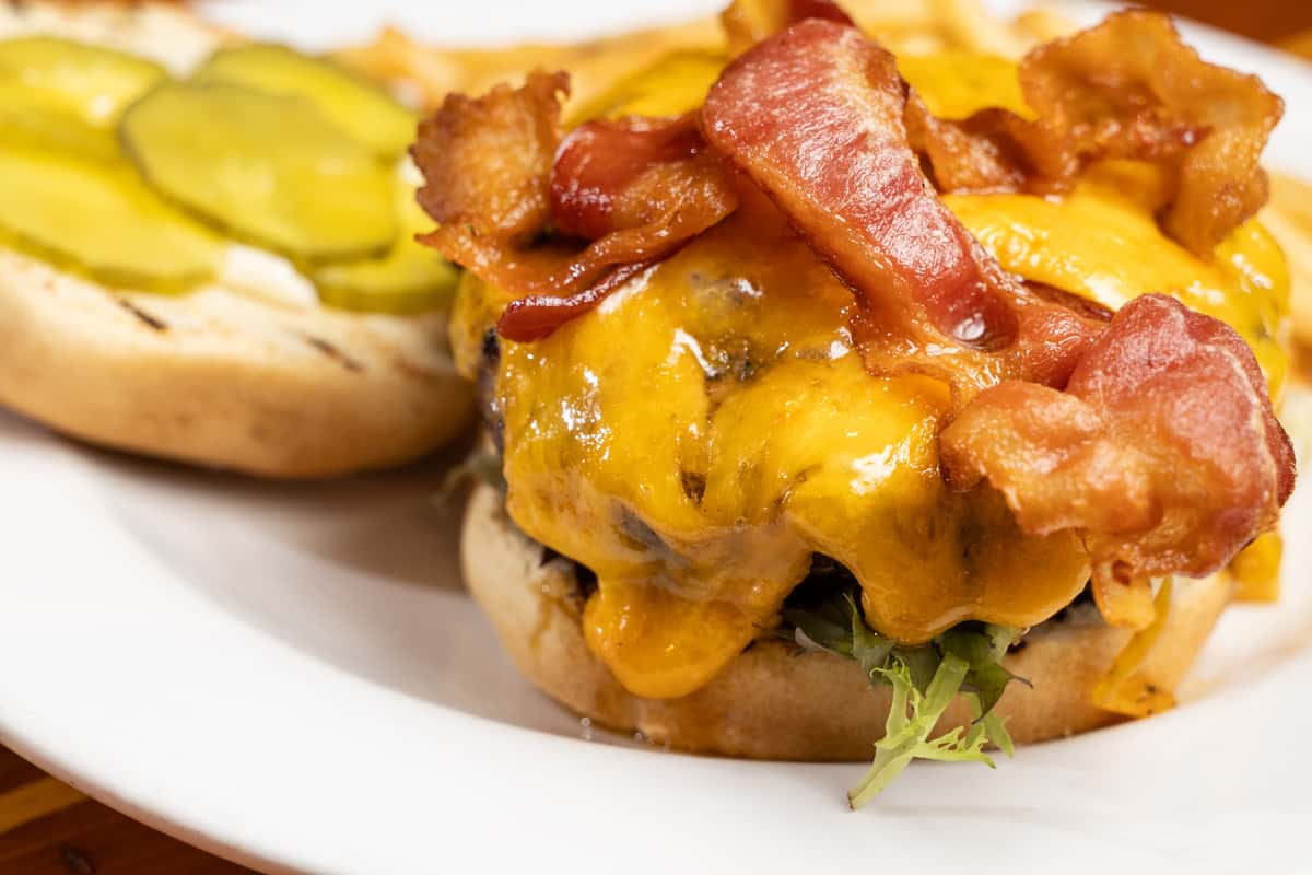 Close-up of a bacon cheeseburger with a juicy beef patty topped with melted cheddar cheese, crispy bacon strips, fresh lettuce, and pickles on a toasted bun, served on a white plate, highlighting classic American bar and grill comfort food.
