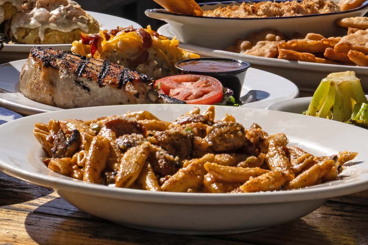 Overhead view of a rustic wooden table filled with assorted American bar and grill dishes, including grilled steak with mashed potatoes, loaded pasta, fried appetizers, roasted vegetables, and shareable plates, showcasing a hearty local bar and grill menu.