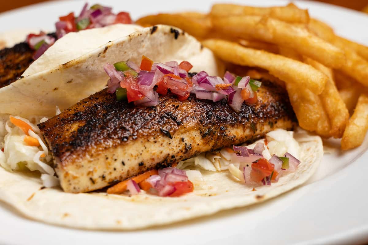 Grilled fish tacos made with seasoned white fish, fresh pico de gallo, and shredded cabbage on soft flour tortillas, served with crispy French fries, showcasing casual seafood bar and grill cuisine.