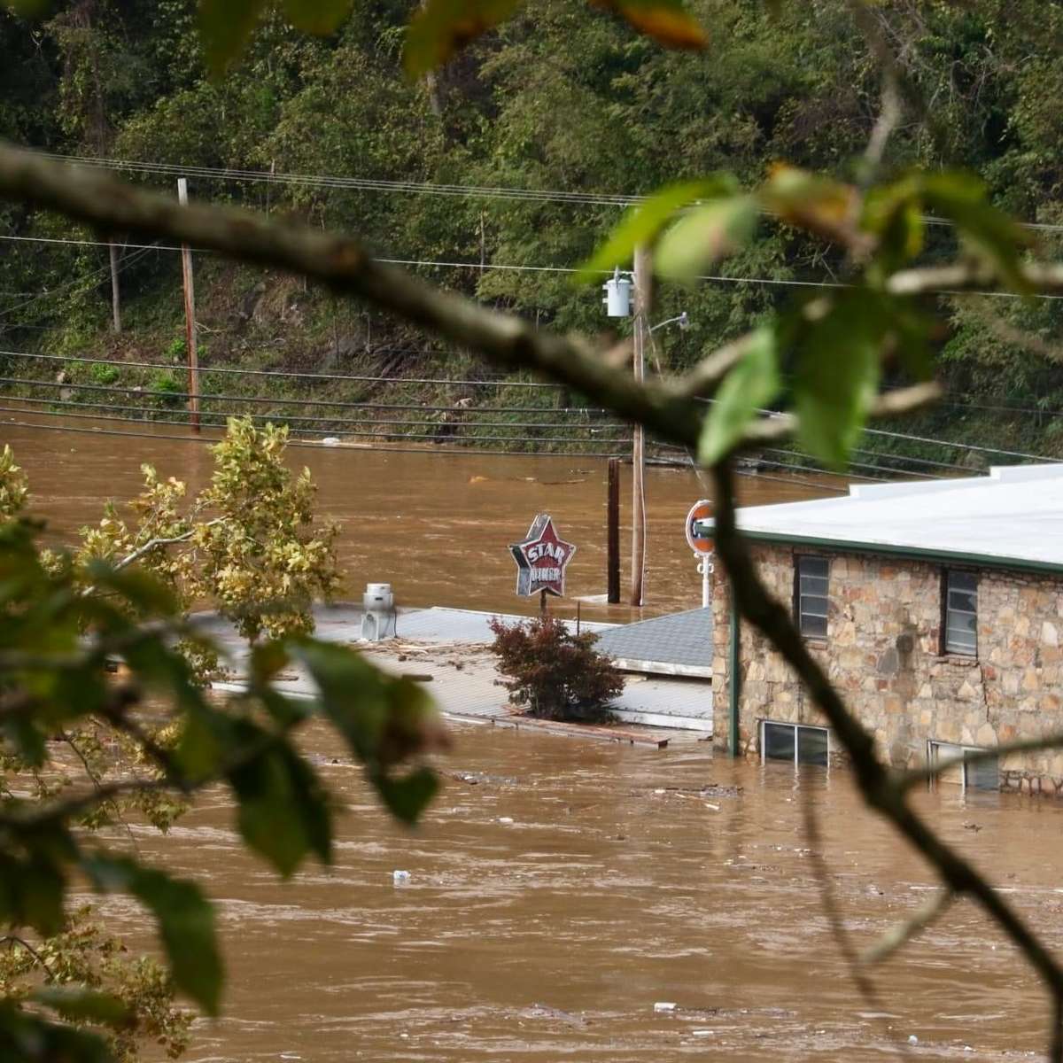 Flood waters surround a building with a 'STAR DINER' sign, viewed through tree branches. The scene depicts the aftermath of severe flooding.
