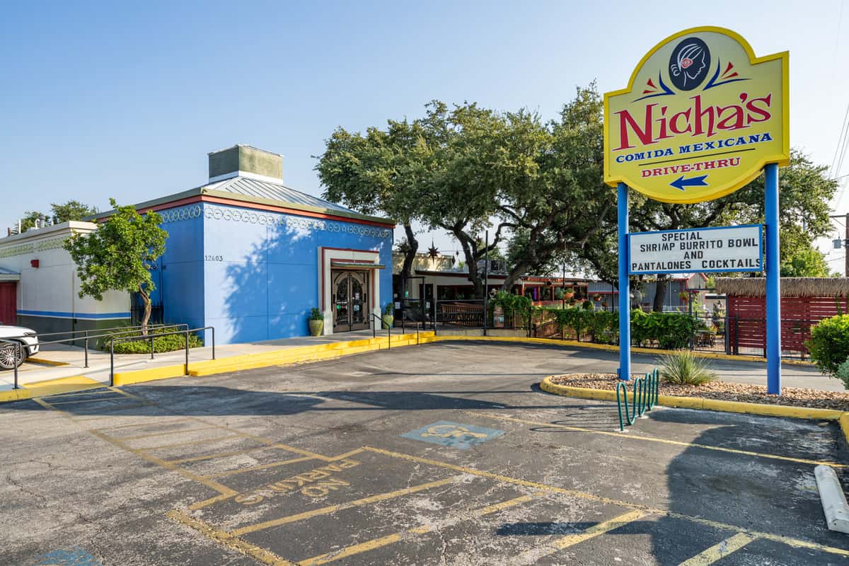 Exterior of Nicha's Comida Mexicana featuring a vibrant blue building, clear signage with drive-thru directions, and visible specials for shrimp burrito bowls and cocktails, set against a backdrop of lush trees and patio seating.