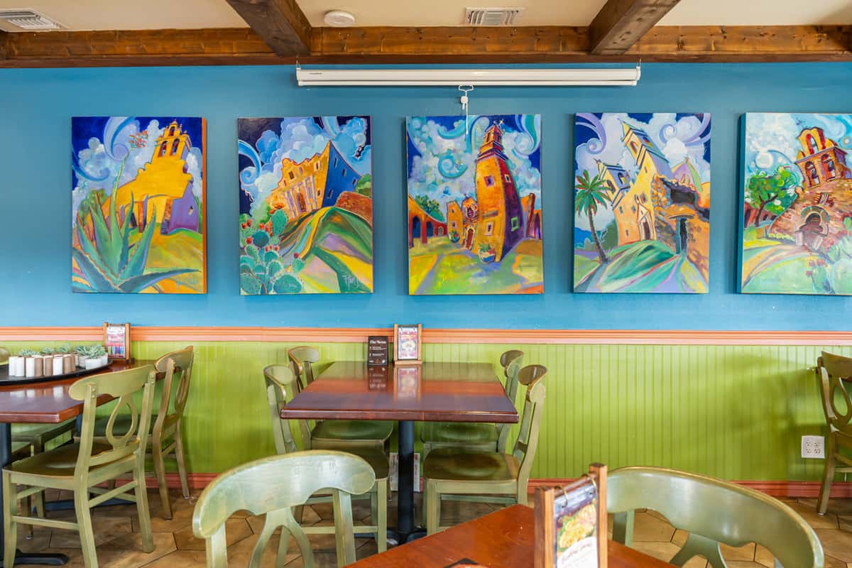 Vibrant dining room wall inside a Mexican restaurant, featuring colorful paintings of mission-style buildings on a bold blue background, paired with green wooden chairs and dark wood tables.
