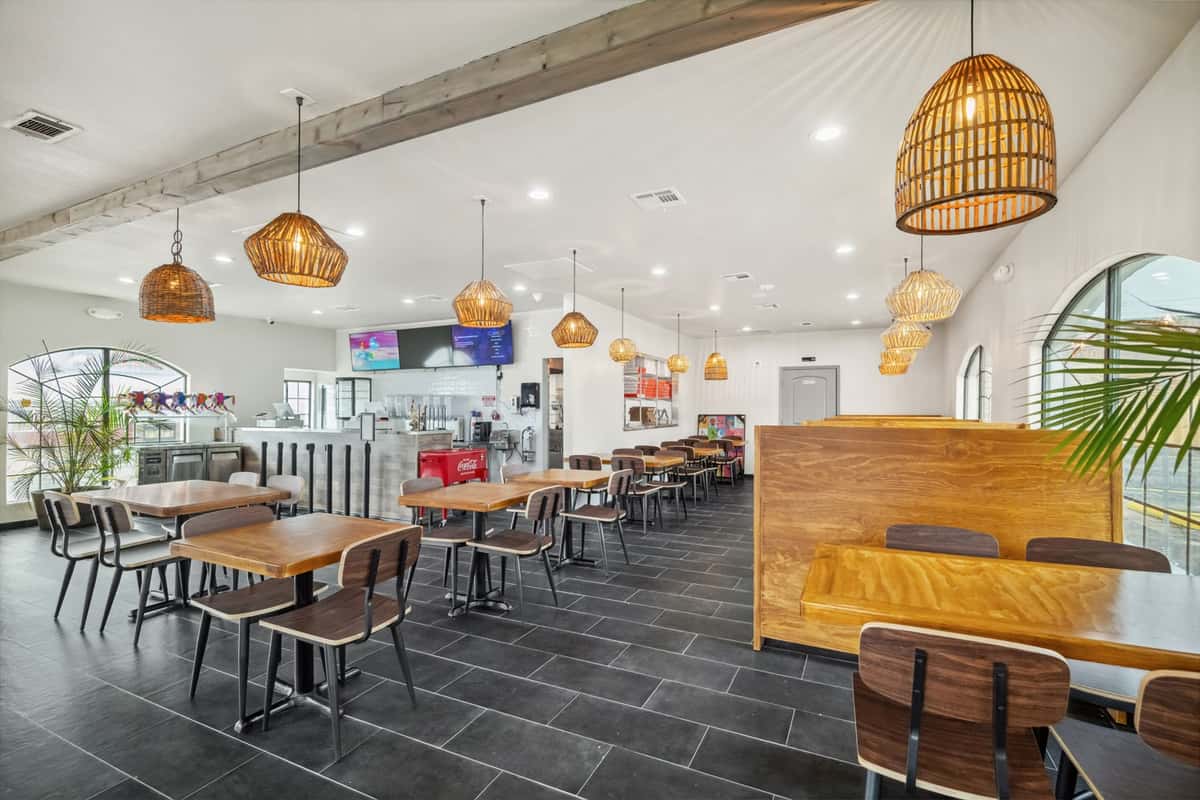 Spacious restaurant dining area with wooden tables and chairs, wicker pendant lights, tiled floor, and visible counter with digital menus.