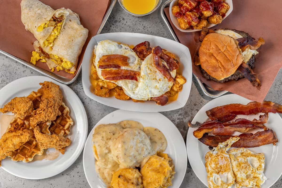Hearty American breakfast spread featuring fried chicken and waffles, bacon and eggs, breakfast burritos, biscuits and gravy, cheesy hashbrowns, and burgers served with tater tots — perfect for diner or brunch restaurant menus.