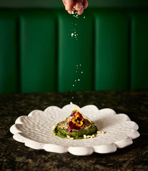 A hand sprinkles seasoning over a beautifully plated Indian dish, featuring vibrant green elements, edible flowers, and pine nuts on a decorative white plate.