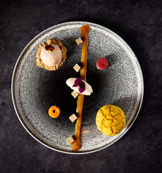 A beautifully plated dessert featuring a scoop of ice cream on a crumb base, drizzled with caramel, surrounded by colorful fruit garnishes and edible flowers, showcasing culinary artistry.