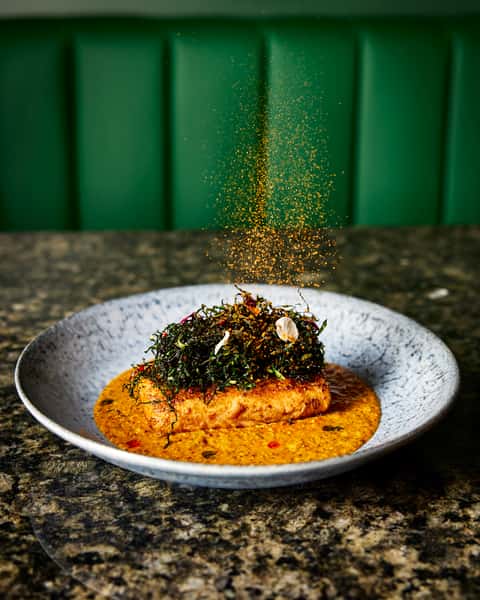 A perfectly cooked salmon fillet topped with crispy greens, served in a rich sauce, with a sprinkle of spices, showcasing the artistry of Indian culinary presentation.
