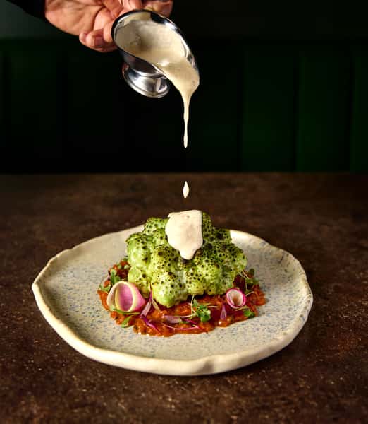 A gourmet dish showcasing a vibrant green cauliflower topped with creamy sauce, garnished with microgreens and radishes, served on a textured plate, highlighting culinary creativity.
