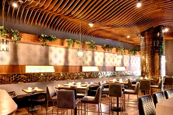 A stylish interior of FITOOR restaurant featuring elegant wooden ceilings, lush greenery, and modern seating arrangements, creating a warm and inviting dining atmosphere.