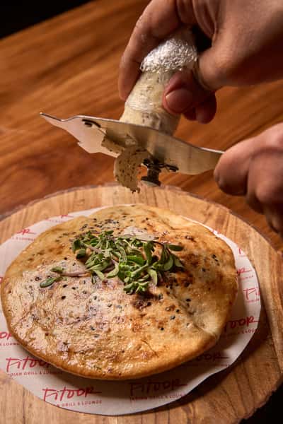 A freshly baked entree, showcasing a golden-brown naan topped with herbs, resting on a wooden board with Fitoor branding, highlighting its delicious presentation.