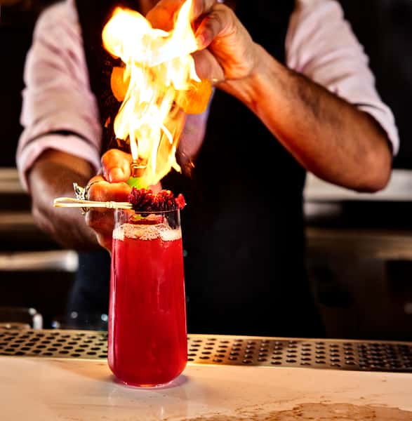 A bartender ignites a cocktail at FITOOR, showcasing a vibrant red drink topped with herbs, creating a dramatic flaming effect that highlights the restaurant's mixology skills.
